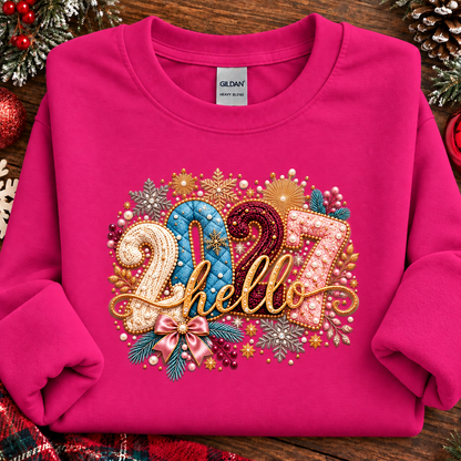 A folded heliconia pink sweatshirt on a rustic wooden surface with holiday decorations around it. The front has a large decorative graphic reading “2027 hello” in gold, blue, pink, and white embellished lettering with snowflakes, stars, and ribbon details.