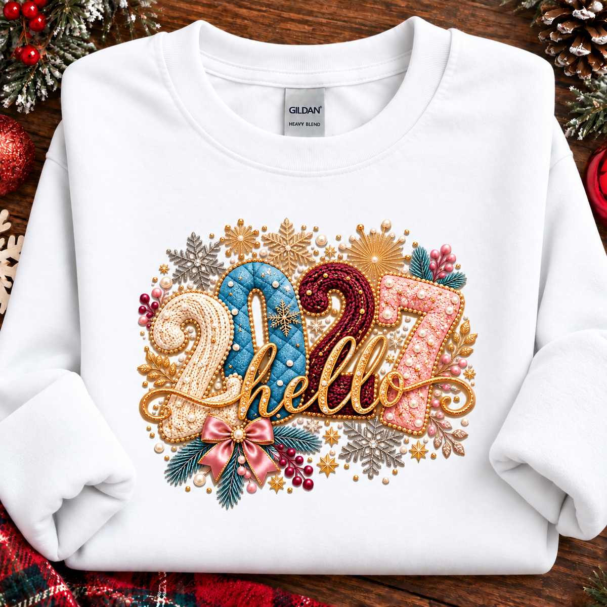 A white Gildan Heavy Weight sweatshirt laid flat on a rustic wooden surface, decorated with a colorful Christmas-themed graphic that reads '2027 hello' in gold, blue, pink, and red tones with festive snowflakes, pinecones, and berries.