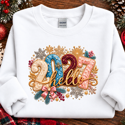 A white Gildan Heavy Weight sweatshirt laid flat on a rustic wooden surface, decorated with a colorful Christmas-themed graphic that reads '2027 hello' in gold, blue, pink, and red tones with festive snowflakes, pinecones, and berries.