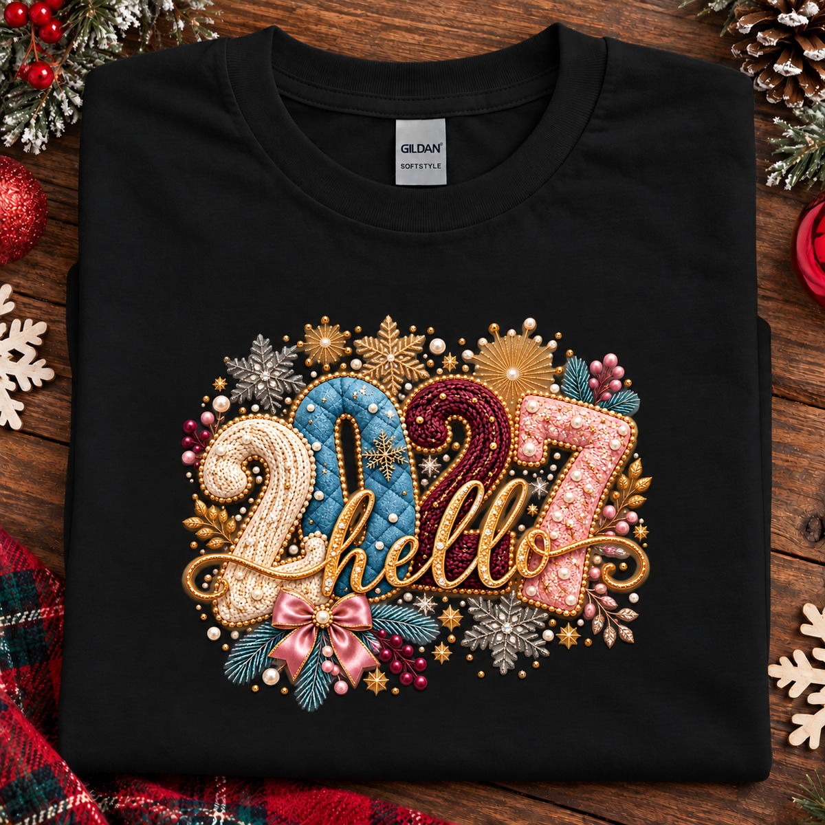 A folded black Gildan t-shirt with a colorful '2027 hello' graphic on the front, styled on a rustic wooden background with Christmas decorations.