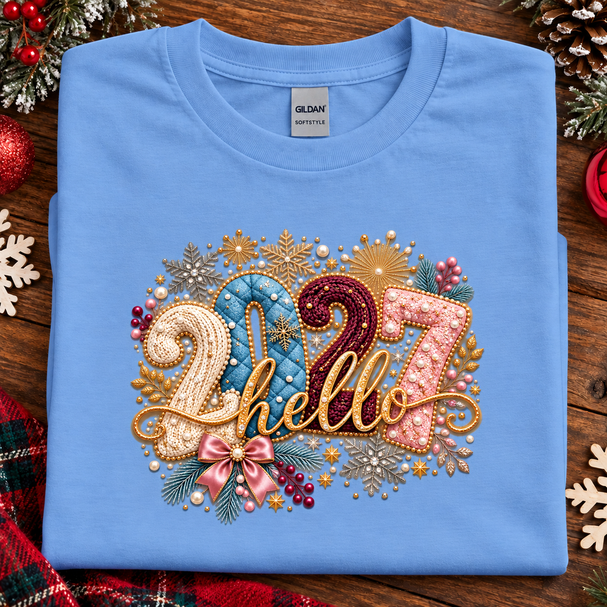 A folded carolina blue Gildan t-shirt with a festive '2027 hello' graphic on the front, styled on a rustic wooden background with holiday decorations.