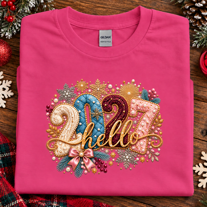 A folded heliconia pink Gildan Botnetka t-shirt with a festive '2027 hello' graphic, styled on a rustic wooden background with Christmas decorations.