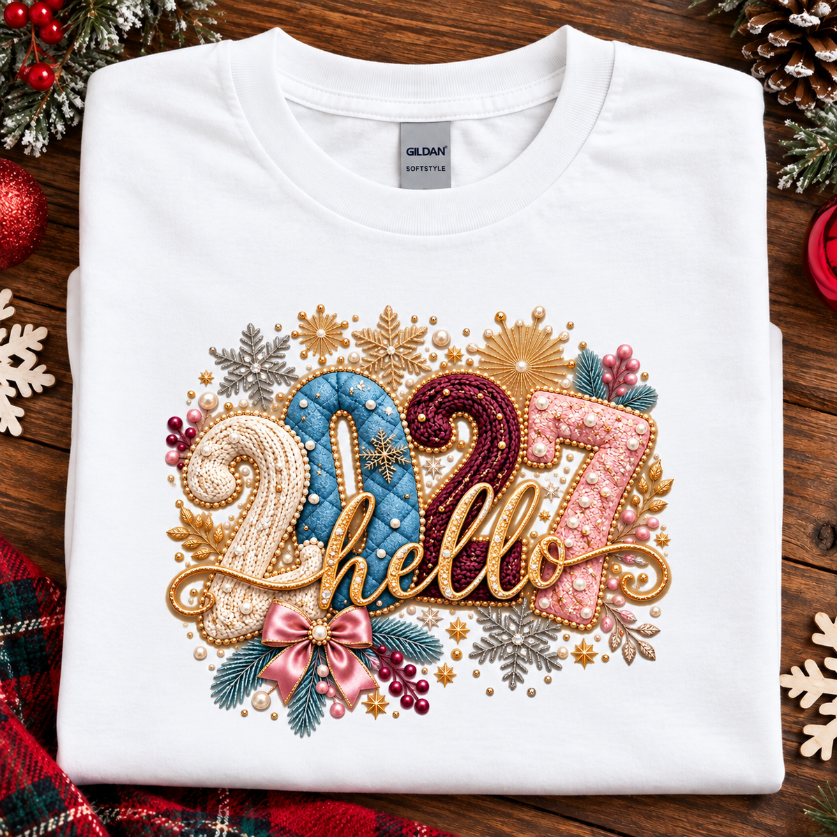 A folded white Gildan Softstyle T-shirt with a large, festive '2027 hello' graphic on the front, styled on a rustic wooden background with holiday decorations like pinecones, a red ornament, and a plaid scarf.