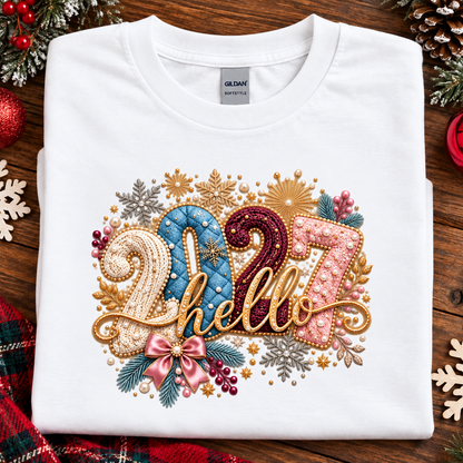 A folded white Gildan Softstyle T-shirt with a large, festive '2027 hello' graphic on the front, styled on a rustic wooden background with holiday decorations like pinecones, a red ornament, and a plaid scarf.