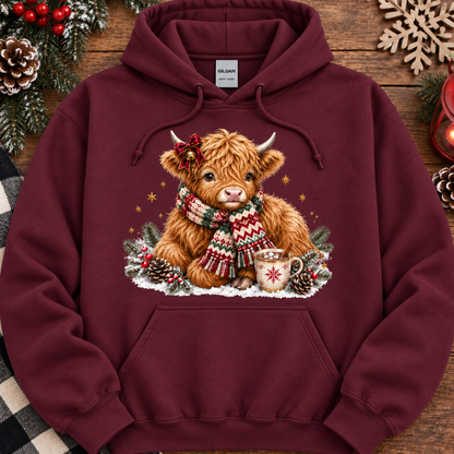 A maroon pullover hoodie laid flat on a rustic wooden surface with holiday decorations around it. The front has a large printed illustration of a fluffy brown Highland cow wearing a red and green knit scarf, surrounded by pinecones, evergreen branches, snow, and small gold stars.