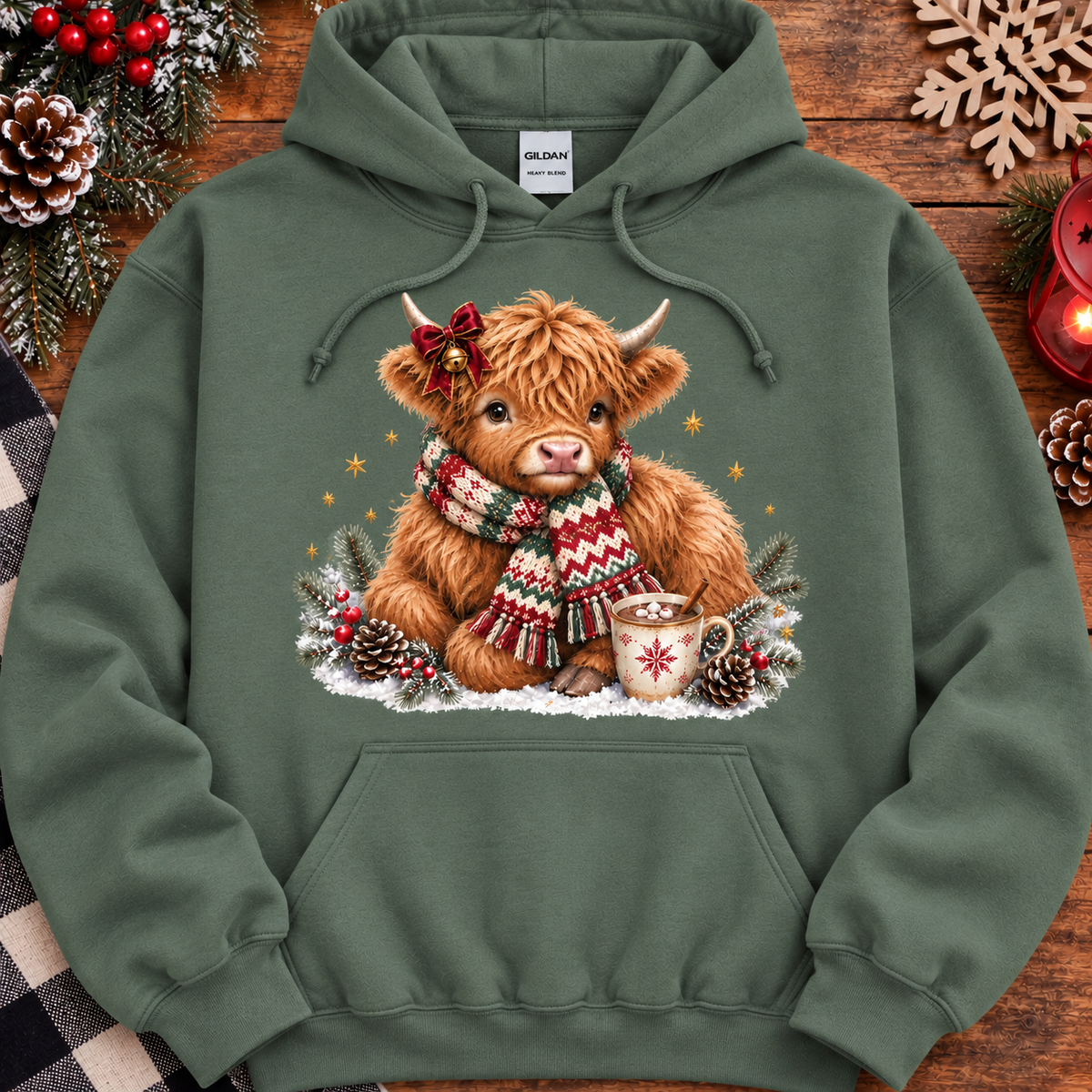 An military green hoodie featuring a festive graphic of a fluffy Highland cow wearing a red and green knit scarf, sitting in the snow with pinecones and a mug. The hoodie is displayed in a flat-lay holiday setting with pinecones, evergreen branches, and a wooden snowflake ornament.