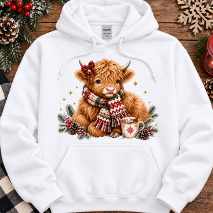 A white pullover hoodie laid flat on a rustic wooden surface, featuring a large front graphic of a fluffy brown Highland cow wearing a red bow and a red-and-white patterned knit scarf, surrounded by pinecones, evergreen branches, and small gold stars. The hoodie includes a drawstring hood and kangaroo pocket.