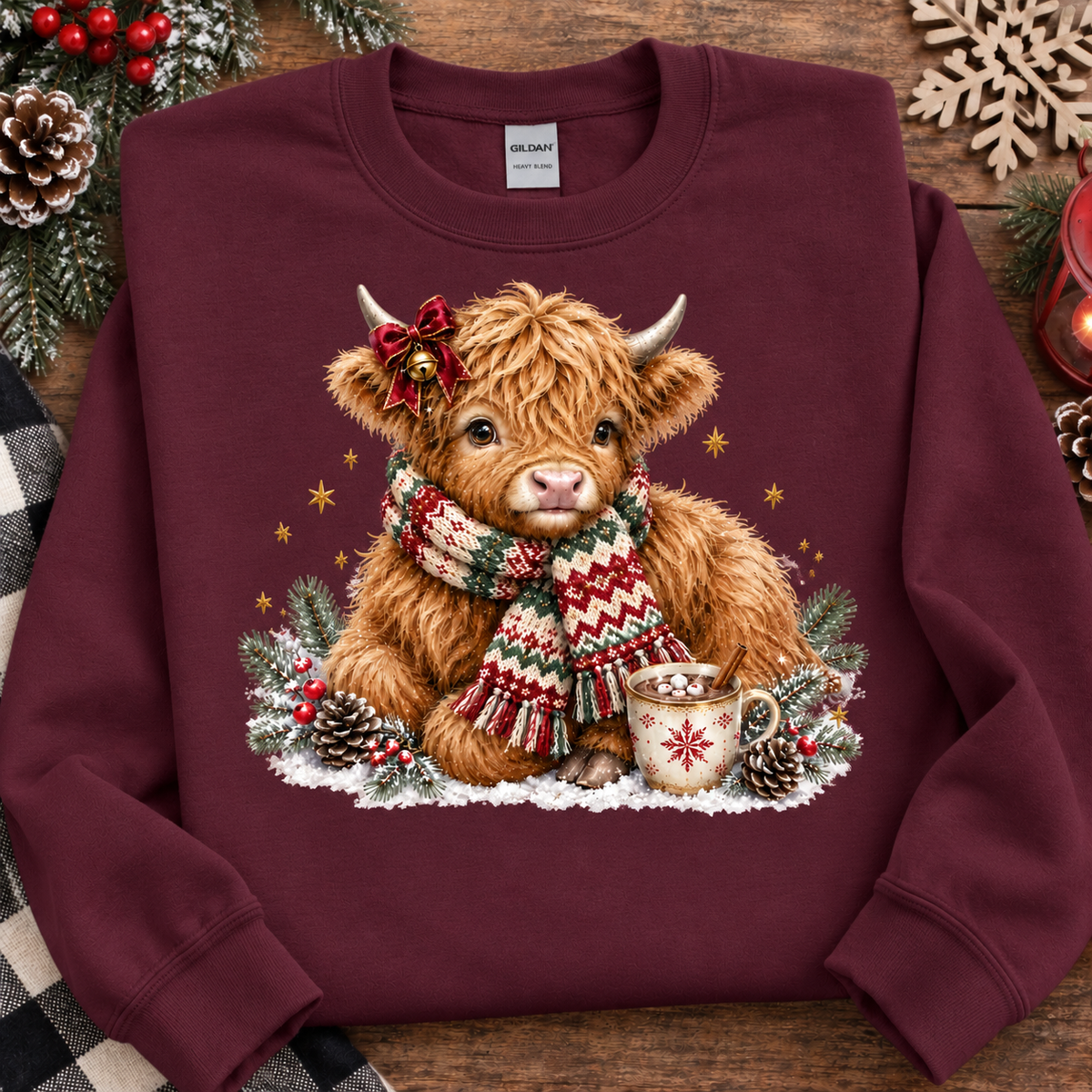 A maroon crewneck sweatshirt laid flat on a rustic wooden surface, decorated with pine branches, pinecones, and red berries. The sweatshirt features a large graphic print of a fluffy Highland cow wearing a red bow and a red, green, and white patterned knit scarf, sitting beside a mug of hot chocolate with snowflakes.