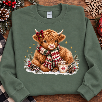 A military green crewneck sweatshirt featuring a festive graphic of a fluffy Highland cow wearing a red bow and a red, green, and white patterned knit scarf, sitting in the snow next to a mug. The sweatshirt is displayed flat on a rustic wooden background decorated with pinecones, evergreen branches, and a wooden snowflake.