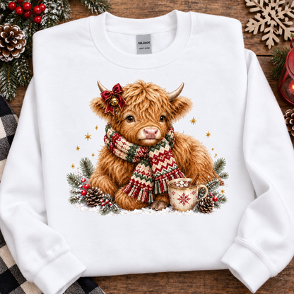 A white crewneck sweatshirt laid flat on a rustic wooden surface, featuring a graphic of a fluffy Highland cow wearing a red bow and a red, green, and white patterned knit scarf, surrounded by pinecones, evergreen branches, and a mug of hot chocolate. The brand Gildan is visible on the inner neck label.