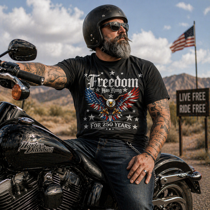 Man wearing a 'Freedom Has Rung For 250 Years' t-shirt sitting on a motorcycle with an American flag in the background.