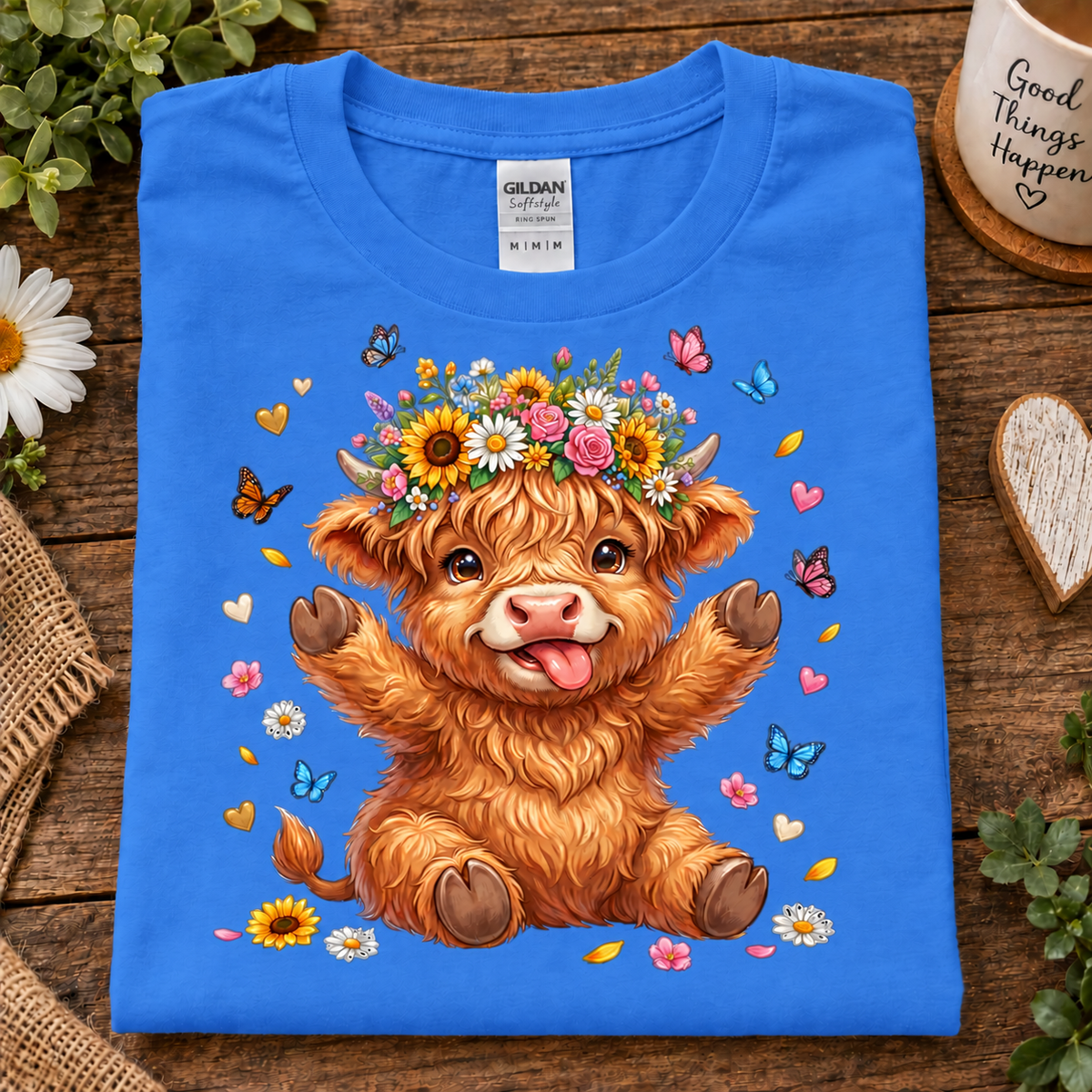 A folded heather royal blue Gildan t-shirt with a cute cartoon Highland cow graphic, displayed on a rustic wooden surface with decorative props.