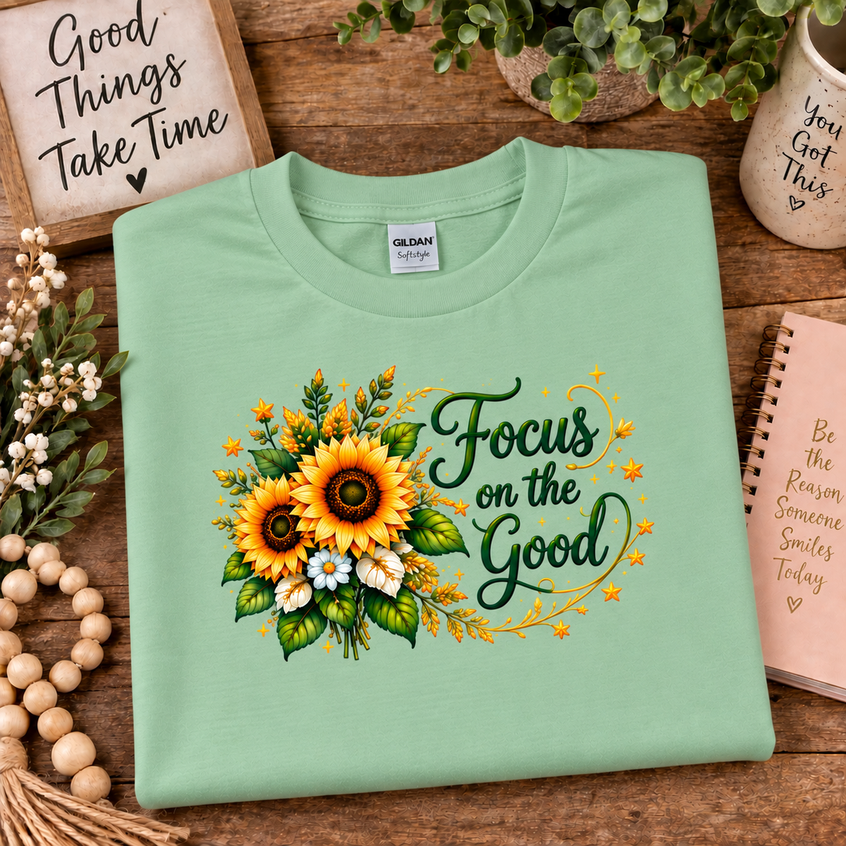 A folded mint green Gildan t-shirt with a sunflower and green script graphic reading 'Focus on the Good', displayed on a rustic wooden table surrounded by decorative props.