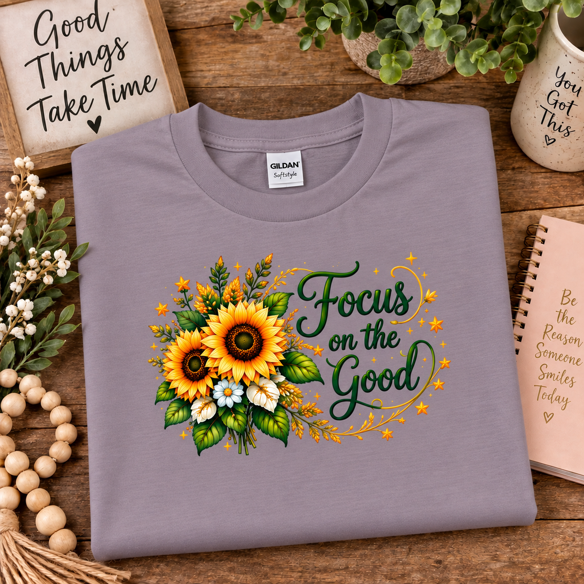 A folded Gildan brand t-shirt in a paragon colored (lavender), featuring a graphic print of two sunflowers, green leaves, and the text 'Focus on the Good' in dark green script. The shirt is styled in a flat-lay arrangement on a rustic wooden table surrounded by decorative props including a wooden bead garland, a sign, a notebook, and a small plant.