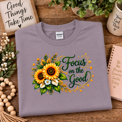 A folded Gildan brand t-shirt in a paragon colored (lavender), featuring a graphic print of two sunflowers, green leaves, and the text 'Focus on the Good' in dark green script. The shirt is styled in a flat-lay arrangement on a rustic wooden table surrounded by decorative props including a wooden bead garland, a sign, a notebook, and a small plant.
