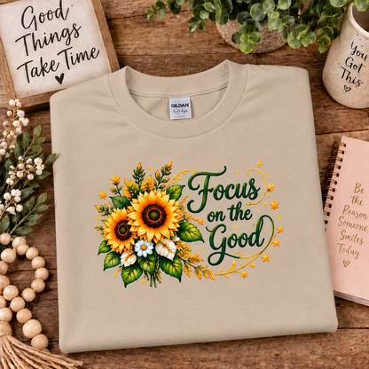 A folded sand colored Gildan t-shirt with a sunflower and green script text graphic reading 'Focus on the Good', styled on a wooden tabletop with decorative props.