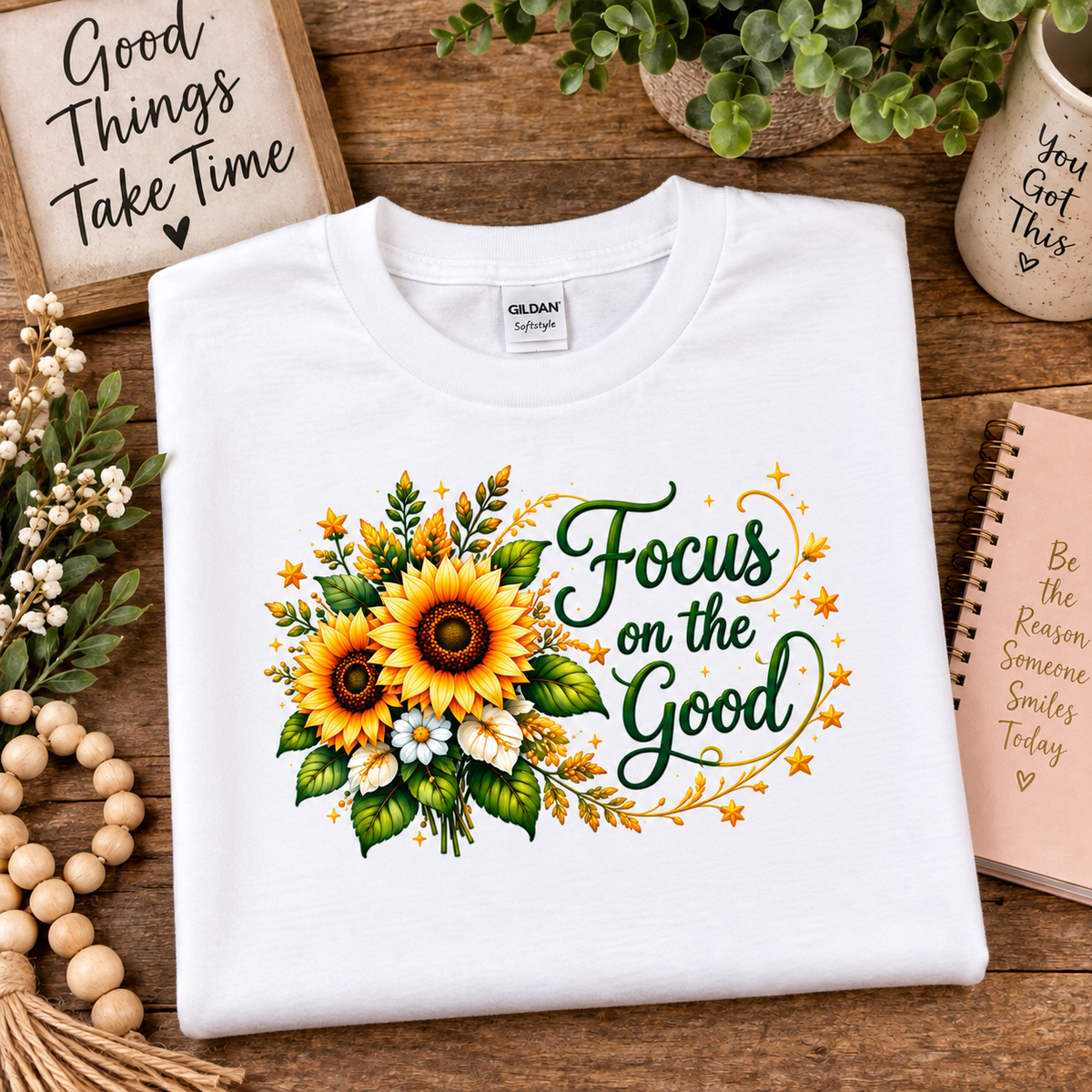A folded white Gildan t-shirt with a sunflower and 'Focus on the Good' graphic, displayed on a wooden tabletop with decorative props.