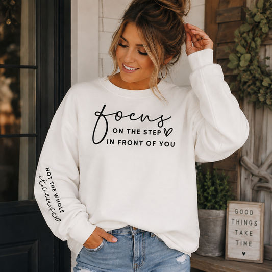 Woman wearing a white sweatshirt with black text Focus on the step in front of you not the whole staircase