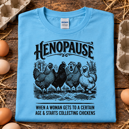 A folded light blue Gildan t-shirt with a humorous henopause graphic, displayed on a rustic wooden background with eggs and cardboard egg cartons.