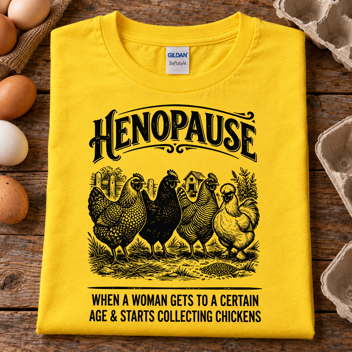 A folded daisy yellow Gildan Softstyle t-shirt with a black graphic print of chickens and the text 'HENOPAUSE WHEN A WOMAN GETS TO A CERTAIN AGE & STARTS COLLECTING CHICKENS', displayed on a rustic wooden surface with eggs and egg cartons around it.