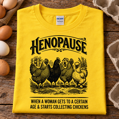 A folded daisy yellow Gildan Softstyle t-shirt with a black graphic print of chickens and the text 'HENOPAUSE WHEN A WOMAN GETS TO A CERTAIN AGE & STARTS COLLECTING CHICKENS', displayed on a rustic wooden surface with eggs and egg cartons around it.