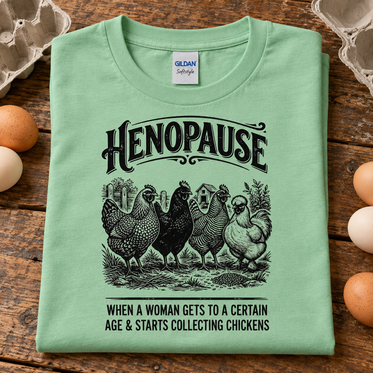 A folded mint light green Gildan Softstyle t-shirt with a humorous hen-themed graphic, displayed on a rustic wooden surface surrounded by eggs.