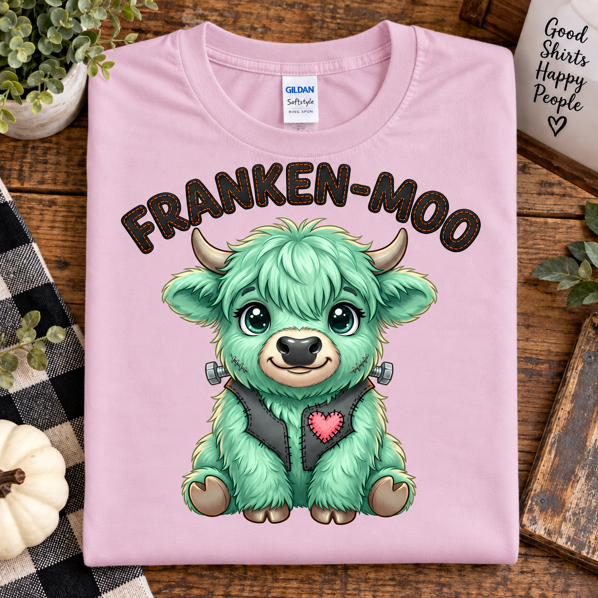 A folded light pink Gildan Softstyle T-shirt with a cartoon monster graphic and the text 'FRANKEN-MOO' on the front, displayed on a rustic wooden surface with decorative plants and props.