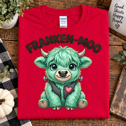 A folded red Gildan Satinway t-shirt with a cartoon graphic of a green highland cow wearing a black vest with a pink heart, with the text 'FRANKEN-MOO' above it. The shirt is displayed on a rustic wooden table surrounded by decorative props including a checkered blanket, a small pumpkin, and a mug.