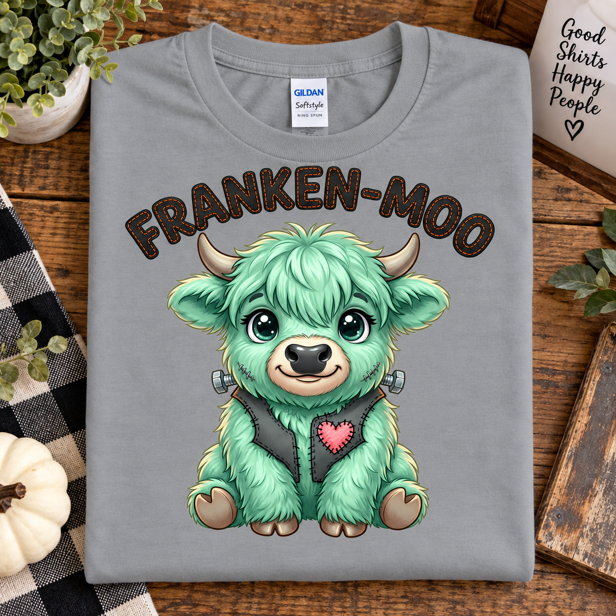 A sport gray Gildan Softstyle t-shirt featuring a cute cartoon Highland cow graphic with the text 'FRANKEN-MOO' above it. The shirt is displayed in a flat-lay setting on a rustic wooden table surrounded by decorative elements like a checkered cloth, a small pumpkin, and a mug.
