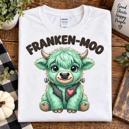 A folded white Gildan Softstyle T-shirt with a colorful cartoon graphic of a green monster calf and the text 'FRANKEN-MOO' printed on the front, styled on a wooden surface with decorative props.