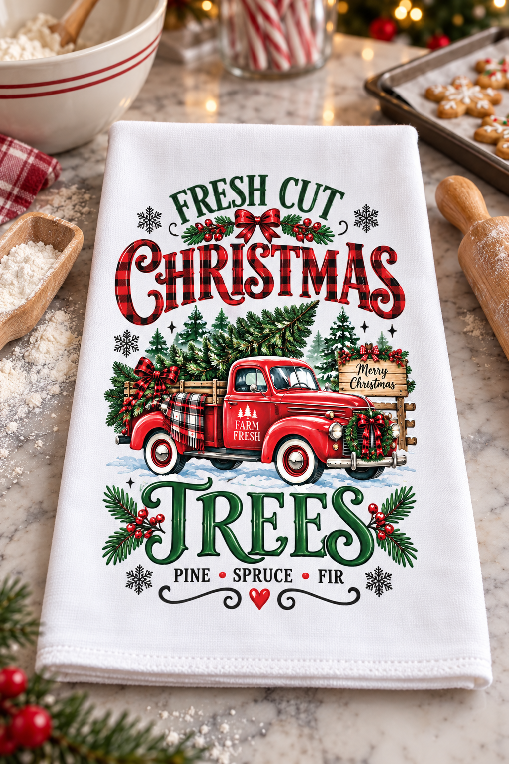 A white kitchen tea towel with a festive Christmas graphic, laid flat on a marble countertop surrounded by holiday cooking props like a rolling pin, a bowl, and a tray of cookies.