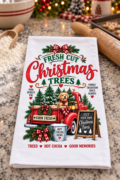 A white kitchen tea towel with a festive Christmas graphic, laid flat on a granite countertop with holiday baking props like a rolling pin, flour, and a mug in the background.