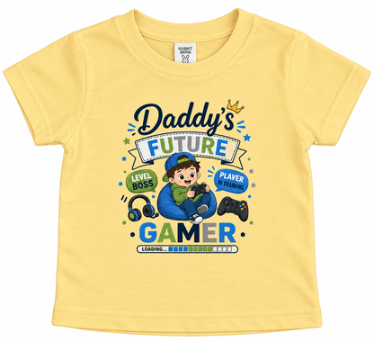 A butter yellow short-sleeve baby t-shirt with a colorful graphic print on the front. The graphic reads 'Daddy's FUTURE GAMER' with a cartoon boy sitting in a blue bean bag, wearing a cap and holding a controller, surrounded by game-themed icons like headphones, a controller, and a crown.