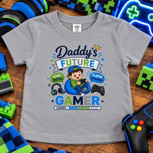 A titanium gray baby t-shirt with a colorful 'Daddy's Future Gamer' graphic, displayed on a wooden surface surrounded by gaming-themed props like controllers, headphones, and pixel blocks.