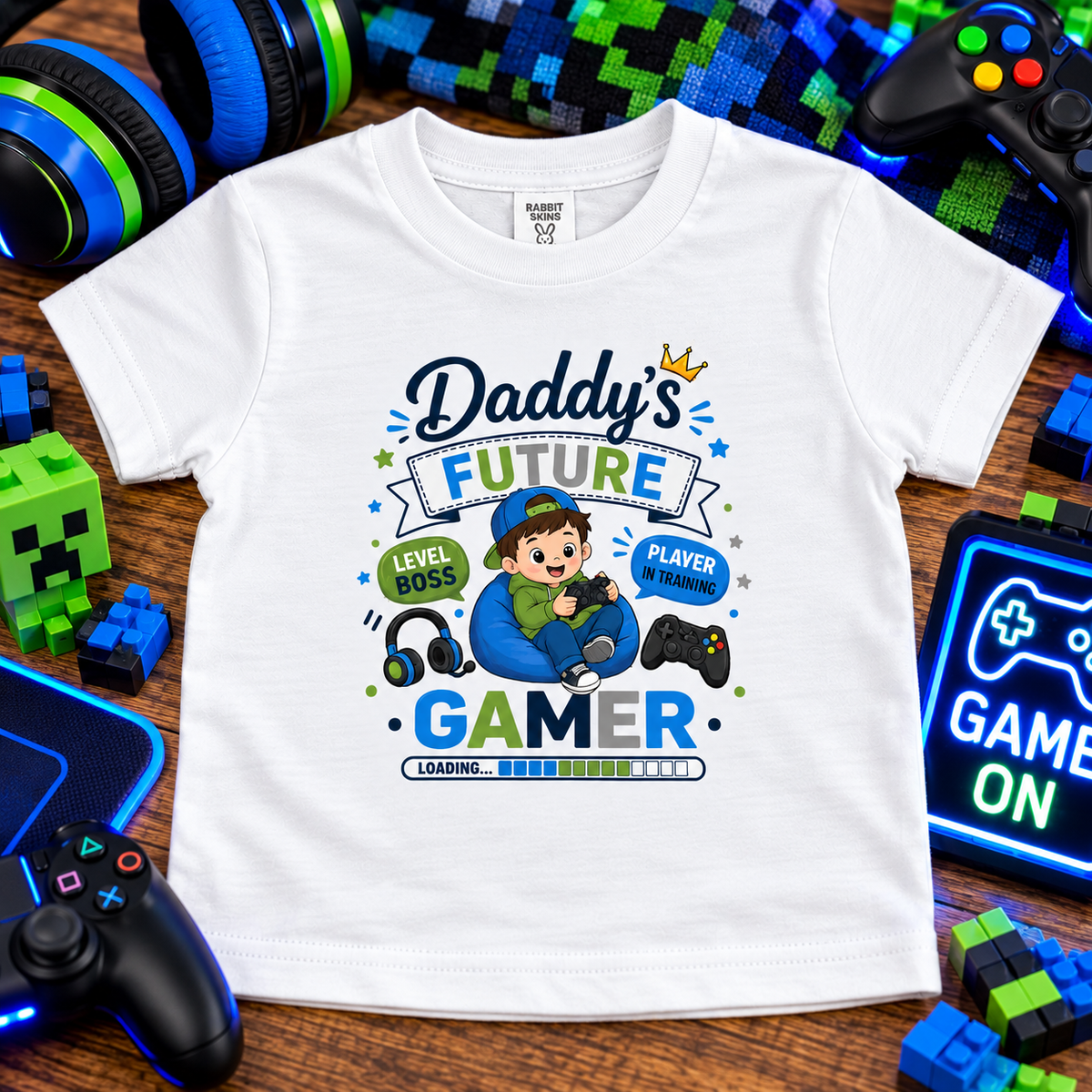 A white short-sleeve graphic T-shirt laid flat on a wooden surface surrounded by gaming-themed props, including controllers, headphones, a pixelated block figure, and a glowing game screen. The shirt graphic reads “Daddy’s Future Gamer” with a cartoon boy, game controller, headset, and progress bar.