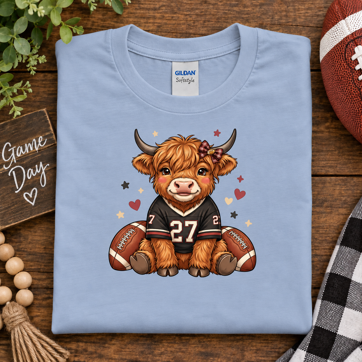 A folded light blue Gildan Softstyle t-shirt is displayed on a rustic wooden surface. The shirt features a cartoon graphic of a fluffy brown Highland cow wearing a black football jersey with the number 27, sitting between two footballs.