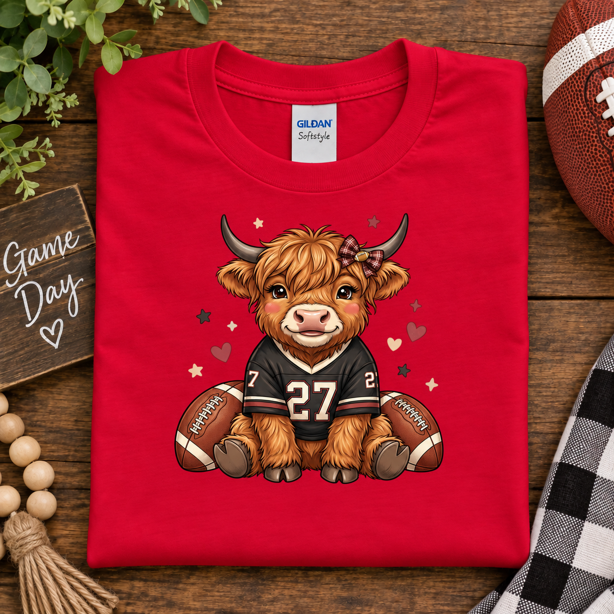 A folded red Gildan Scenery t-shirt is displayed on a rustic wooden table. The shirt features a cartoon graphic of a fluffy Highland cow wearing a football jersey with the number 27, sitting between two footballs with small hearts and stars scattered around it.