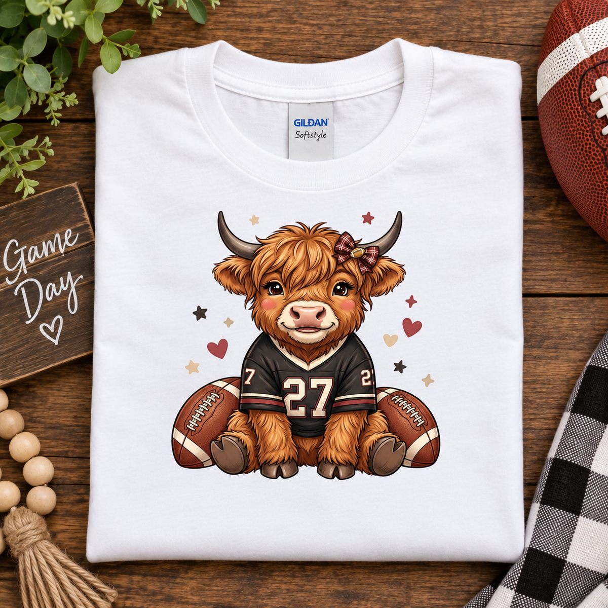 A folded white Gildan Softstyle t-shirt is displayed on a rustic wooden background with football-themed props, including a football, wooden beads, a plaid cloth, and a 'Game Day' sign. The shirt features a cartoon graphic of a Scottish highland cow wearing a black football jersey with the number 27.