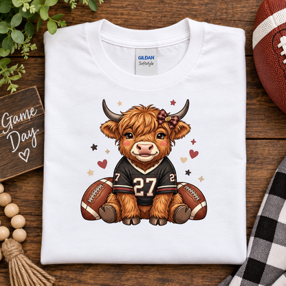 A folded white Gildan Softstyle t-shirt is displayed on a rustic wooden background with football-themed props, including a football, wooden beads, a plaid cloth, and a 'Game Day' sign. The shirt features a cartoon graphic of a Scottish highland cow wearing a black football jersey with the number 27.