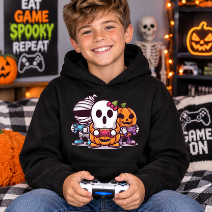 A smiling young boy wearing a black hoodie with a colorful cartoon graphic of a ghost, mummy, and pumpkin holding a game controller. He is sitting on a couch in a cozy, decorated room with Halloween and gaming themed decor.