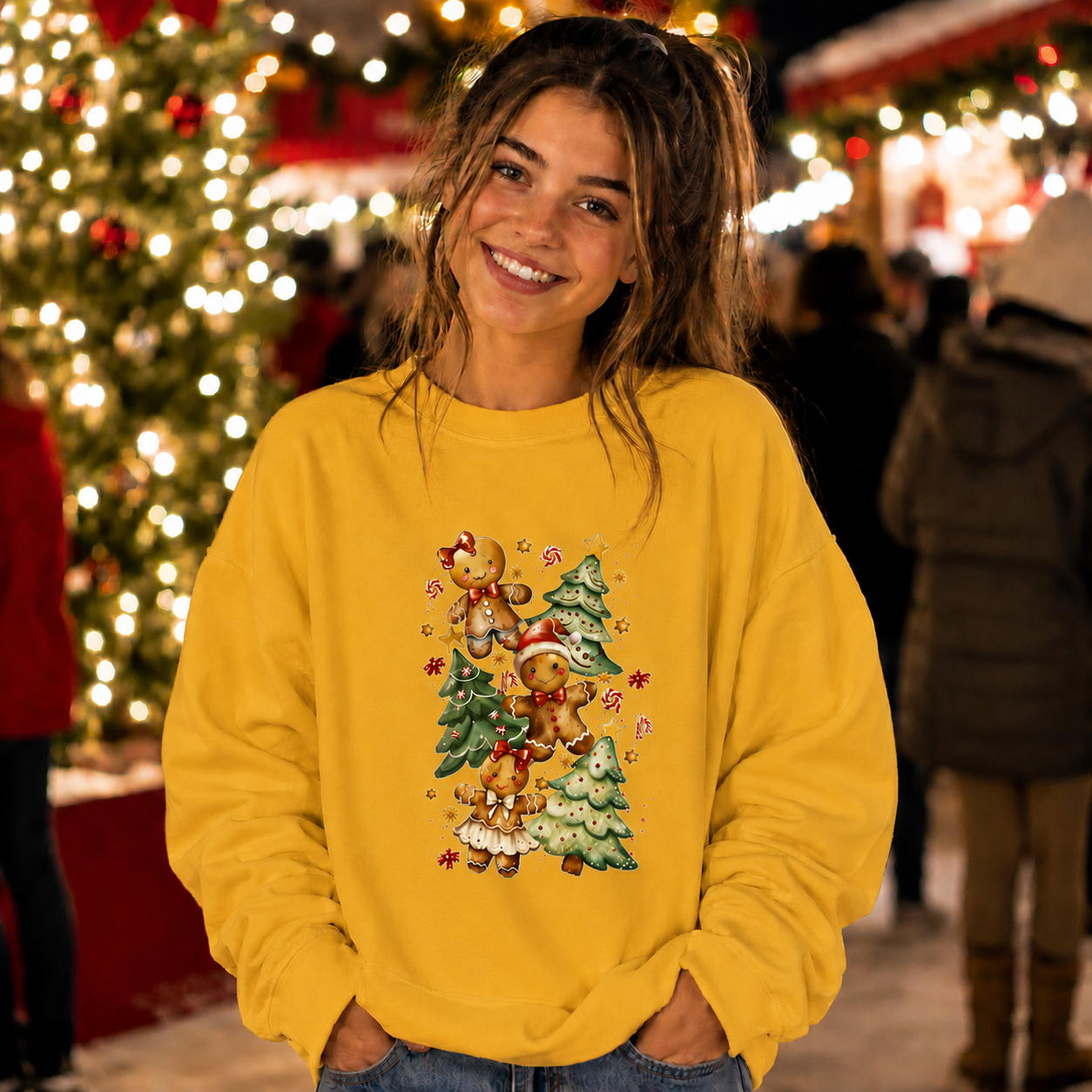 A woman smiling in a festive holiday market, wearing a bright yellow crewneck sweatshirt with a colorful graphic of teddy bears, a Christmas tree, and candy canes. The background is filled with warm, glowing Christmas tree lights.