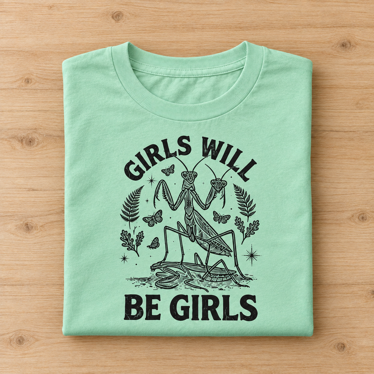 A folded mint green t-shirt with a black graphic print of a praying mantis and the text 'GIRLS WILL BE GIRLS', displayed flat on a light wood background.