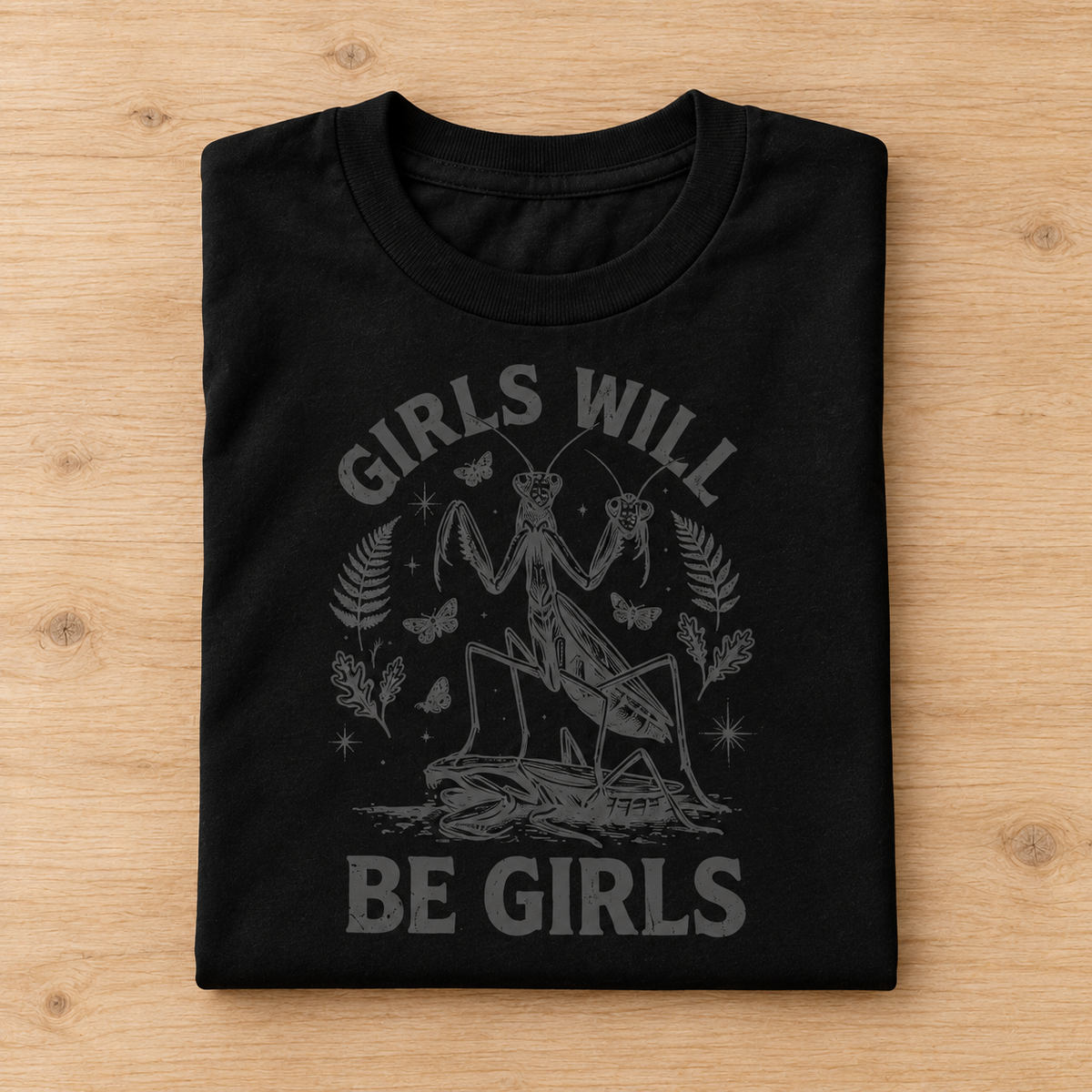 A folded black short-sleeve T-shirt with a dark gray graphic print on a light wood background. The graphic features a praying mantis, butterflies, stars, and the text 'GIRLS WILL BE GIRLS'.