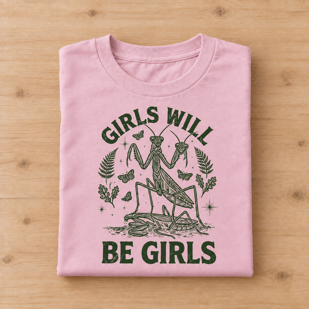 A folded light pink t-shirt with a dark green graphic of a praying mantis and the text 'GIRLS WILL BE GIRLS', displayed flat on a light wood background.