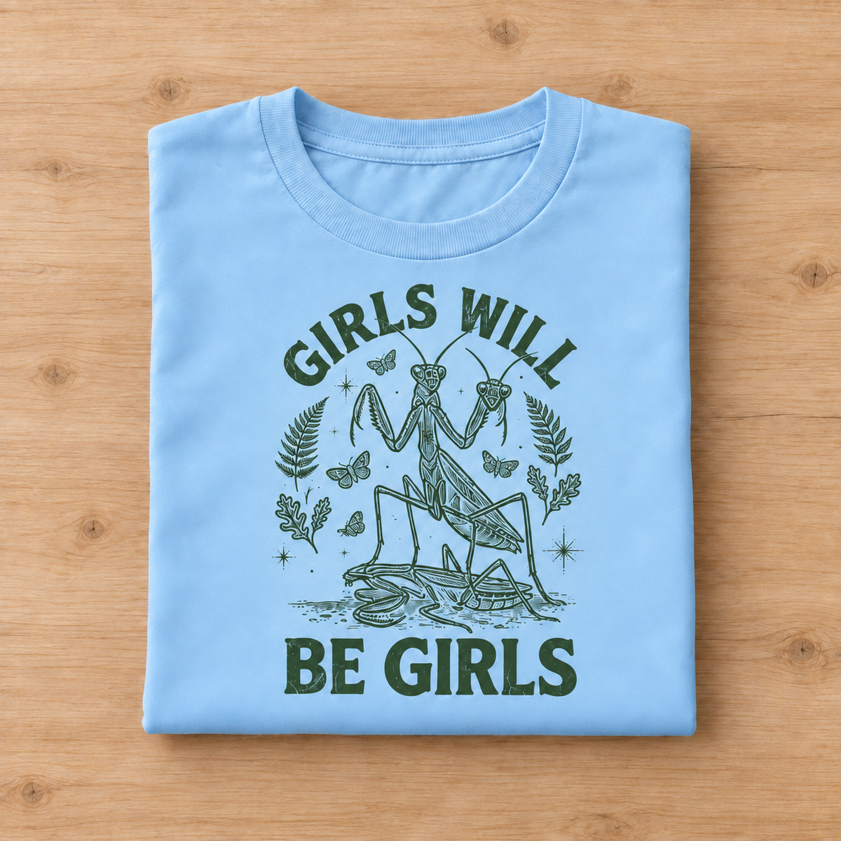 A folded sky blue t-shirt with a dark green graphic print of a praying mantis and butterflies, featuring the text 'GIRLS WILL BE GIRLS', displayed flat on a light wood background.