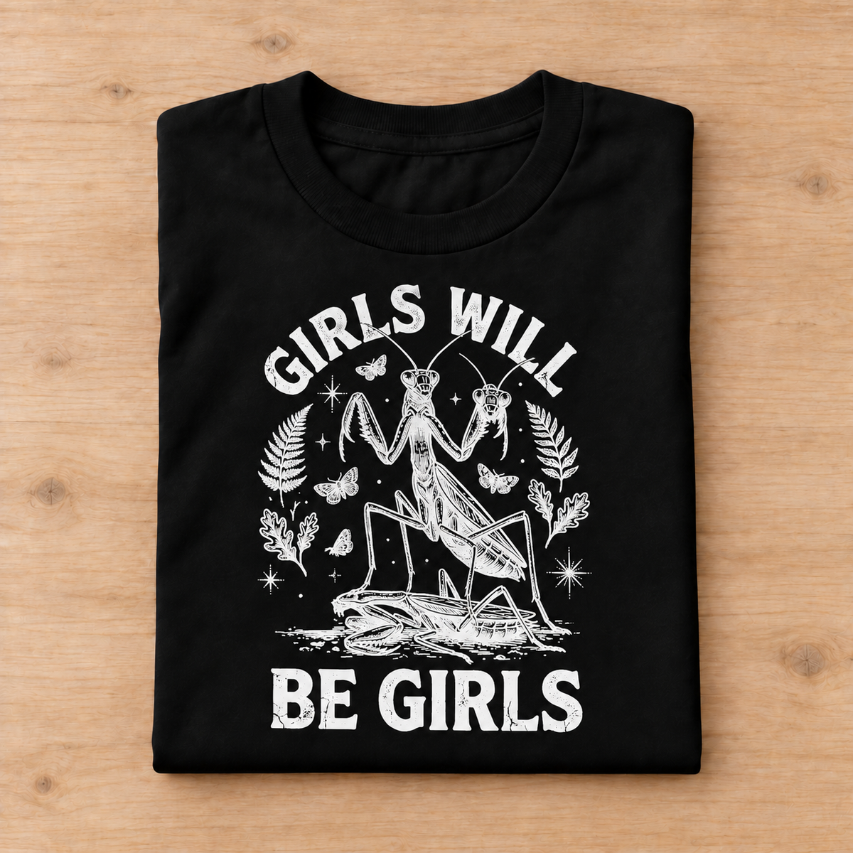 A folded black t-shirt with a white graphic print of a praying mantis and the text 'GIRLS WILL BE GIRLS', displayed flat on a light wood background.