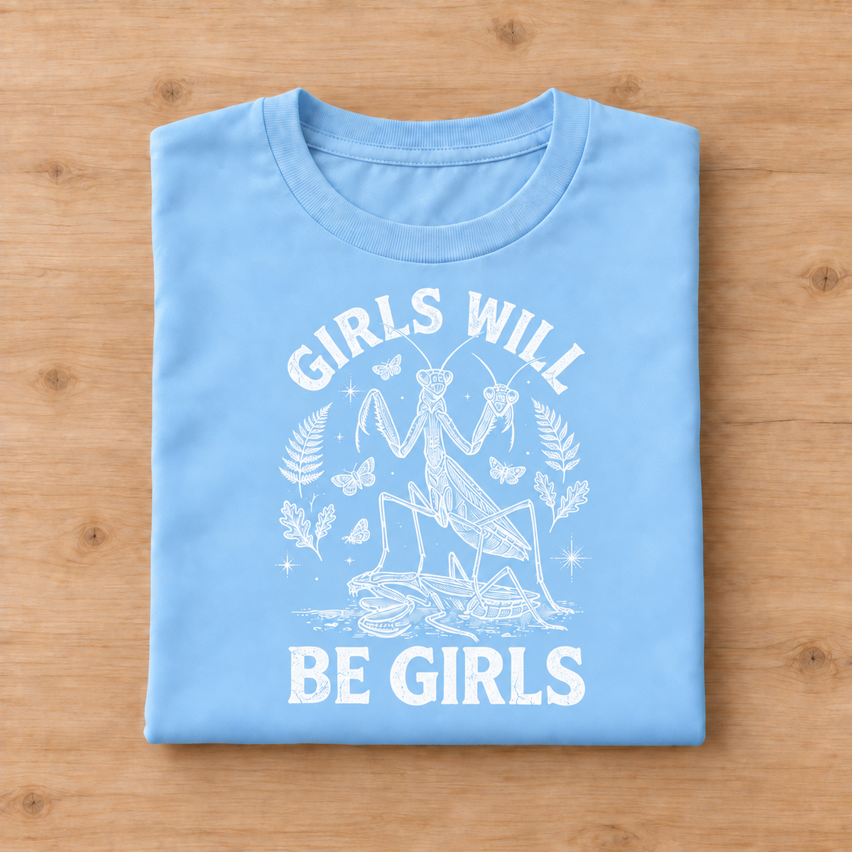 A folded light blue t-shirt with a white graphic print of a praying mantis and the text 'GIRLS WILL BE GIRLS', displayed flat on a light brown wooden surface.