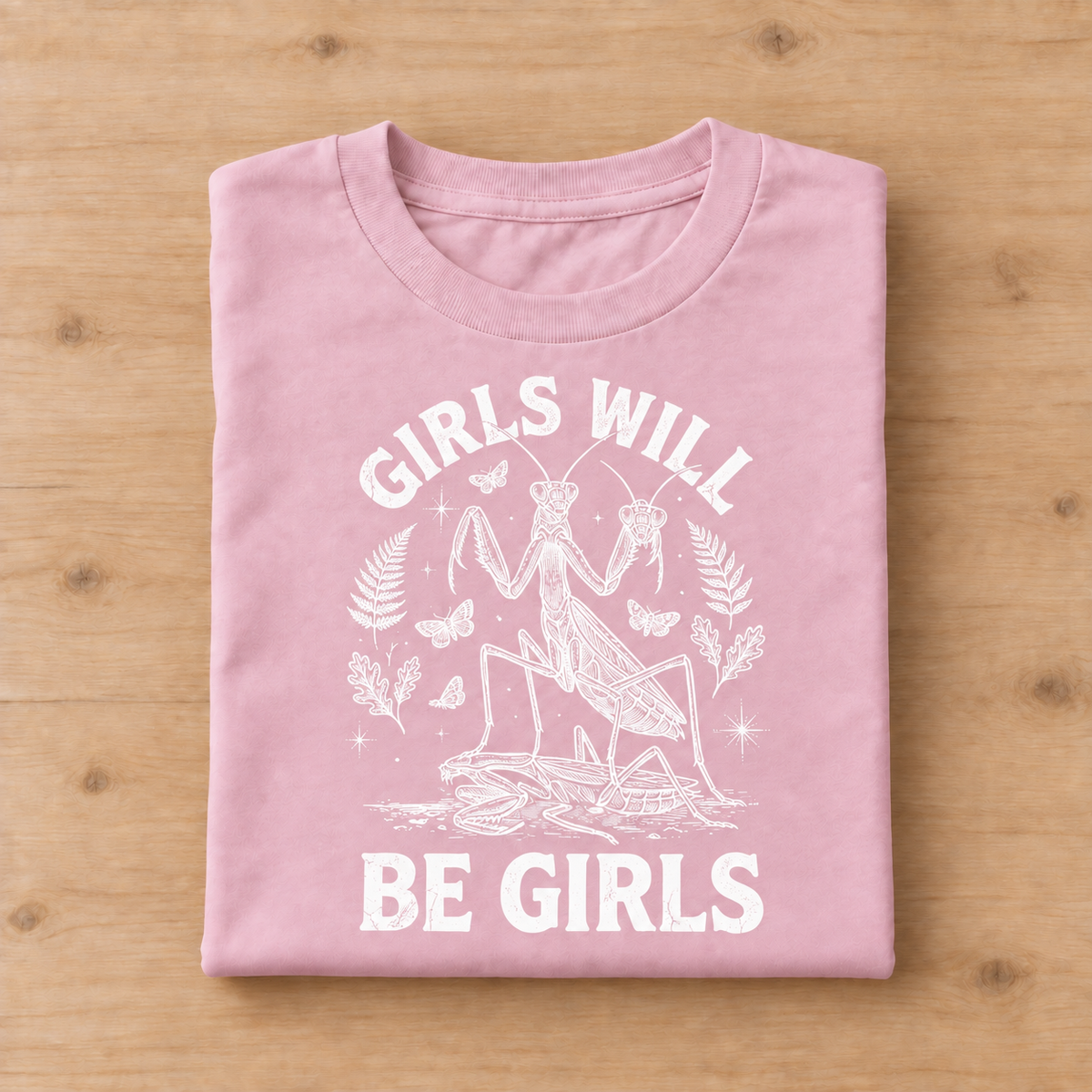A folded light pink t-shirt with a white graphic print of a praying mantis and the text 'GIRLS WILL BE GIRLS', displayed flat on a light wood background.