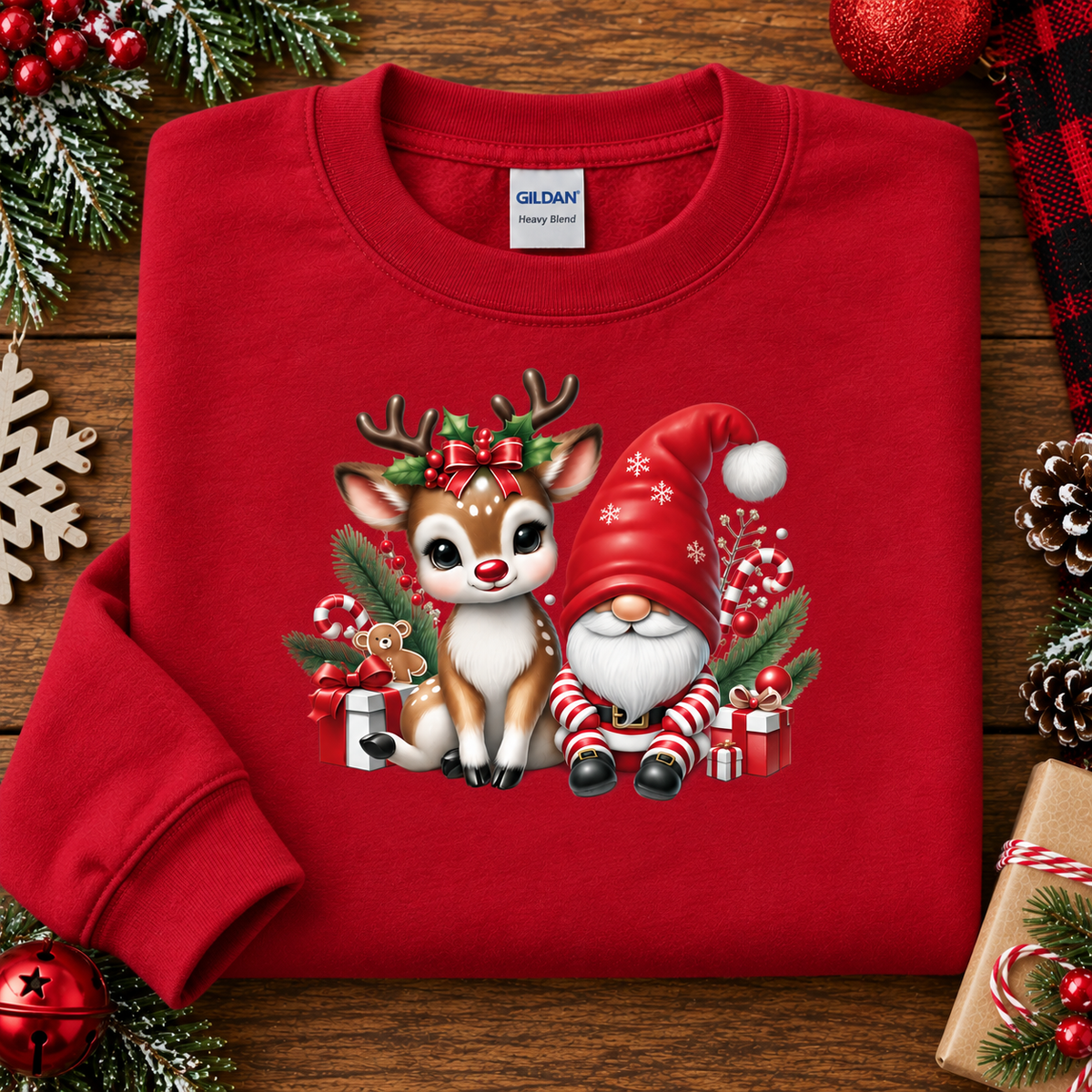 A folded red crewneck sweatshirt with a Christmas graphic of a cute white and brown reindeer and a Santa-clad gnome, styled on a wooden surface with holiday decorations.