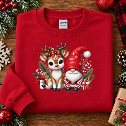 A folded red crewneck sweatshirt with a Christmas graphic of a cute white and brown reindeer and a Santa-clad gnome, styled on a wooden surface with holiday decorations.